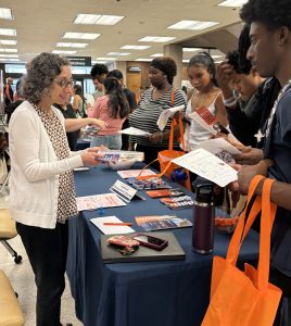 Students standing at table, listening to and taking fliers and paper from a staff person.