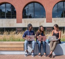 In front of the Schine Student Center students sit with laptops and laugh.