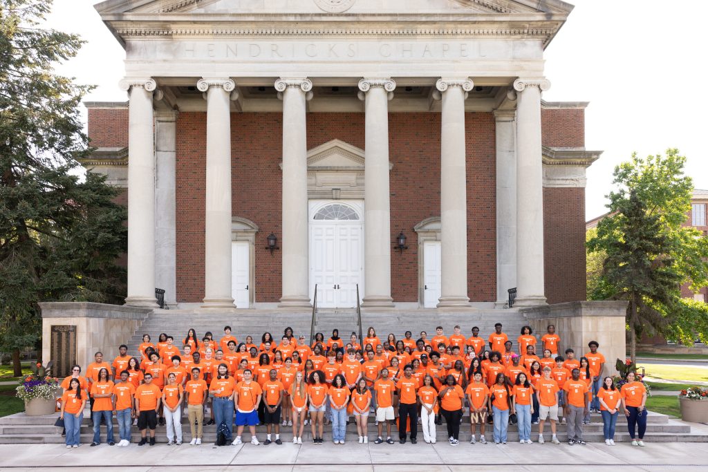Group of students in SummerStart t-shirts standing together, with Hendricks Chapel building in background.